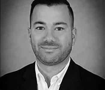 White male in front of grey/black background with black suit jacket and white button up shirt